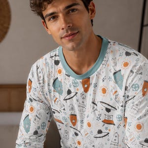 Puede incluir: Una camiseta de manga larga con cuello redondo en color verde azulado apagado. La prenda presenta un estampado integral sobre fondo blanco compuesto por pequeños soles naranjas, ilustraciones espaciales en verde azulado y naranja, y ovnis.