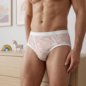 May include: Briefs featuring a light pink background with a repeating pattern of white unicorns, colourful rainbows, and small hearts. The underwear has a solid white elastic waistband and white trim along the leg openings and front panel seams.