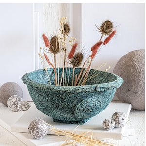May include: A textured teal bowl with a rough, uneven rim and a circular embossed geometric design on the side. The bowl contains a collection of dried brown teasel seed heads and slender, reddish-brown grass plumes arranged upright.