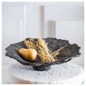Puede incluir: Un cuenco decorativo gris oscuro y texturizado con base de pedestal. El cuenco contiene una pera y tallos de trigo secos. Una pieza única de decoración del hogar.