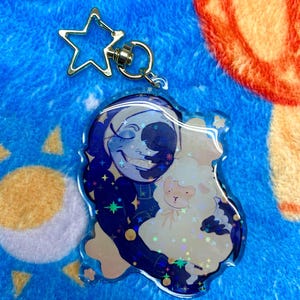 May include: A clear acrylic keychain featuring a character with a dark blue moon-themed face and starry clothing, holding a small, fluffy white sheep. The keychain has a silver-colored star-shaped clasp and a holographic finish with star-shaped sparkles.
