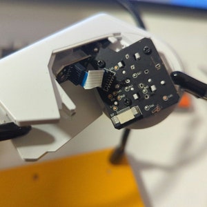 May include: A close-up of a white electronic device with a circuit board visible. The circuit board has a black background with white text and components. The device is held in place by two black clamps.
