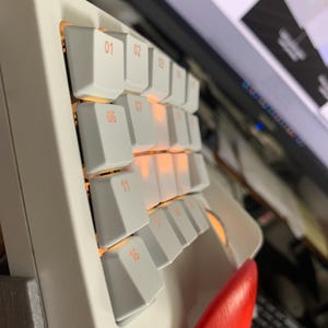 May include: A white mechanical macro pad featuring a grid of keys with orange backlighting. Each keycap is labeled with a numerical identifier, including 01, 02, 03, 06, 07, 11, 16, and 17. A bright red wrist rest is attached to the bottom of the device.