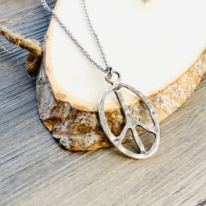 May include: A silver peace sign pendant necklace with a hammered finish. The necklace is on a silver chain and is displayed on a wooden surface.