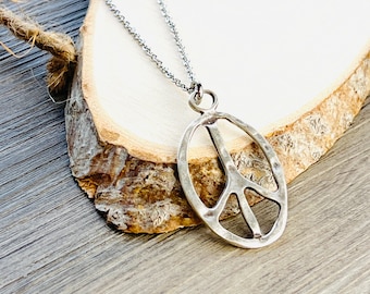 May include: A silver peace sign pendant necklace with a hammered finish. The necklace is on a silver chain and is displayed on a wooden surface.