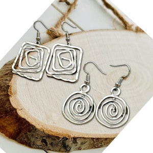 May include: Two pairs of silver-colored earrings. One pair features a square design with a layered, open-frame pattern. The other pair has a circular, spiral design. Both pairs have hook closures and are displayed on a wooden surface.