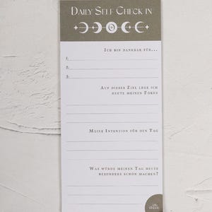 Op de afbeelding: Een 'Daily Self Check In' notitieblok met een grijze header met maanfasesymbolen. Het notitieblok heeft lijnen voor schrijfopdrachten zoals 'Ik ben dankbaar voor...' en 'Mijn intentie voor de dag'.