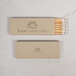 Può includere: Due scatole di fiammiferi beige. La scatola superiore è aperta, rivelando fiammiferi con punte bianche. La scatola superiore ha un logo a forma di sole e la scritta "Shine Your Light". La scatola inferiore ha il logo "Oh Shanti" e l'indirizzo del sito web.