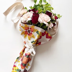 May include: A bouquet of flowers wrapped in a fabric with a quilt pattern, featuring red, pink, and white flowers, and green foliage. The bouquet is held by a cream-colored strap.