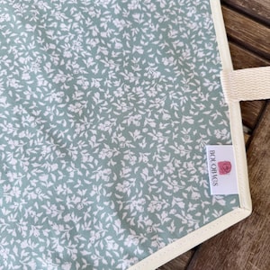 Może przedstawiać: Artykuł z tkaniny z turkusowym tłem i białym wzorem kwiatowym. Tekst "White floral on teal" jest nadrukowany w lewym g&oacute;rnym rogu. Biała metka z tkaniny z napisem "BOLOBAGS" i ikoną r&oacute;żowej r&oacute;ży jest przymocowana z boku.