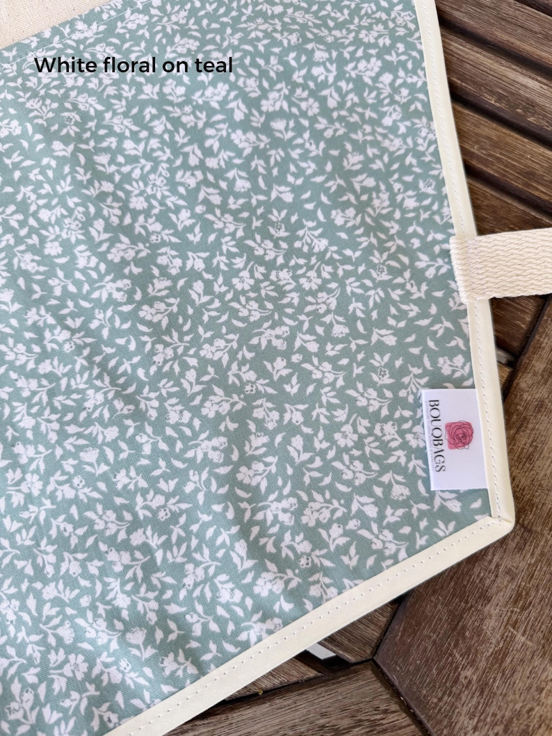 Może przedstawiać: Artykuł z tkaniny z turkusowym tłem i białym wzorem kwiatowym. Tekst "White floral on teal" jest nadrukowany w lewym g&oacute;rnym rogu. Biała metka z tkaniny z napisem "BOLOBAGS" i ikoną r&oacute;żowej r&oacute;ży jest przymocowana z boku.