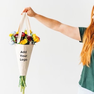 May include: A cone-shaped beige canvas flower bag with two fabric handles, featuring the black printed text Add Your Logo on the front. The bag is filled with a colorful bouquet of yellow sunflowers, red roses, and blue flowers with green stems.