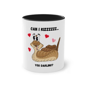 Könnte beinhalten: Weiße Keramik-Tasse mit schwarzem Innenrand. Die Tasse zeigt eine Cartoon-Schlange mit dem Text "CAN I KIZZZZZZ... YOU DARLING?" und Herzillustrationen. Die Schlange ist braun und hellbraun.