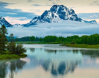 Puede incluir: Paisaje panorámico con un río tranquilo que refleja montañas nevadas. Frondosos árboles verdes bordean las orillas, con un cielo nublado de fondo. La escena evoca una sensación de tranquilidad y belleza natural.