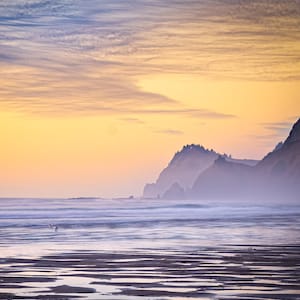 May include: A scenic landscape photograph of a beach at sunset. The sky transitions from yellow to blue, with wispy clouds. Mountains are silhouetted in the distance, and the wet sand reflects the colors of the sky.