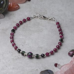 May include: A bracelet made with pink and black beads, with a larger green and purple bead in the centre. The bracelet has a silver clasp.