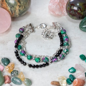 May include: A multi-strand bracelet with black, green, and pink beads, silver chain links, and a silver charm in the shape of a crescent moon with a tree design.