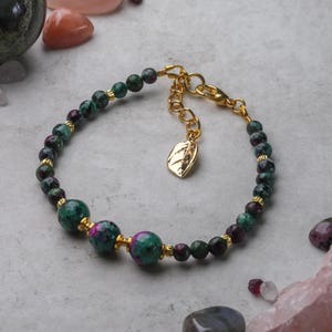 May include: A green and pink gemstone bracelet with gold beads and a gold leaf charm. The bracelet is made with small round beads and two larger round beads.