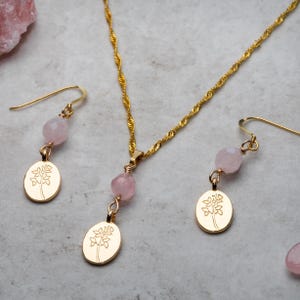 May include: A gold-coloured jewellery set comprising a necklace and earrings. The necklace features a delicate chain with a pendant of a pink gemstone and a gold-coloured oval charm with a floral design. The earrings have a similar design.