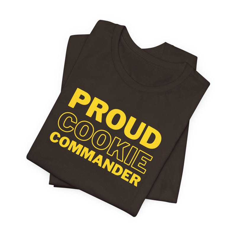 May include: A brown t-shirt with the text "PROUD COOKIE COMMANDER" in yellow.