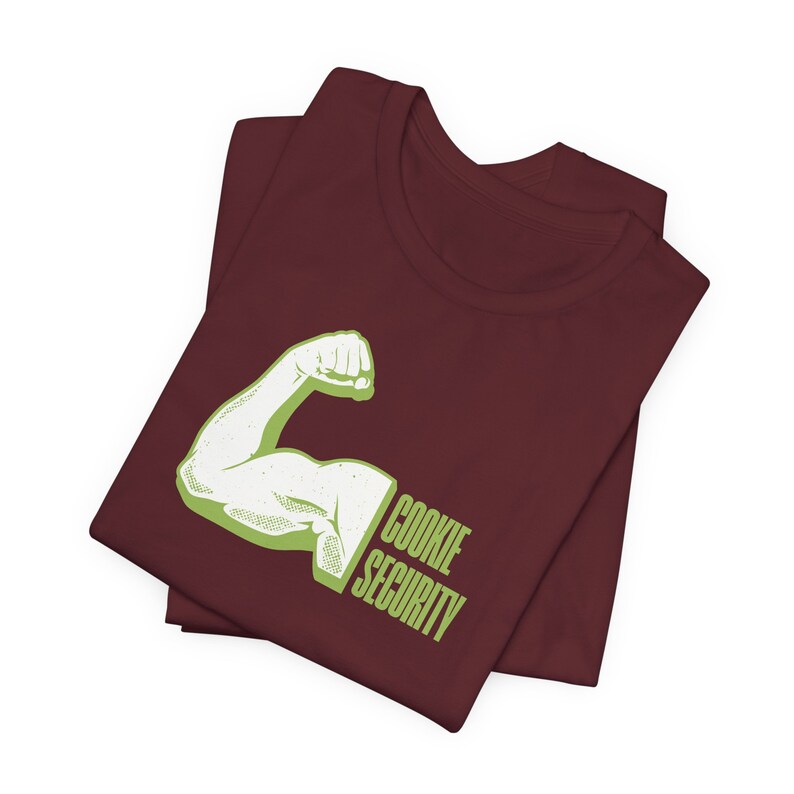 Puede incluir: Una camiseta marr&oacute;n con un gr&aacute;fico verde de un b&iacute;ceps flexionado con el texto "COOKIE SECURITY" debajo.