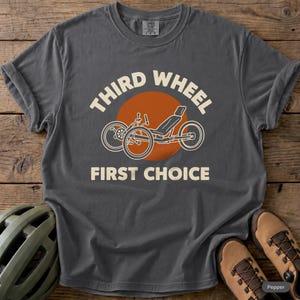 May include: A charcoal gray t-shirt featuring a graphic of a recumbent tricycle centered over a burnt orange circle. The text "THIRD WHEEL" is arched above the graphic, and "FIRST CHOICE" is printed below in a cream-colored sans-serif font.