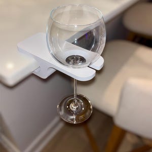 Puede incluir: Una copa de vino transparente descansa en un soporte blanco para copas de vino con clip. El soporte está sujeto al borde de una superficie blanca. El tallo de la copa de vino es visible y la copa está vacía.