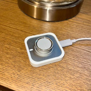 May include: A white and silver wireless charging pad for a smartwatch. The pad has a square shape with a silver center and a white border. A white charging cable is connected to the pad. The pad is on a wooden surface.