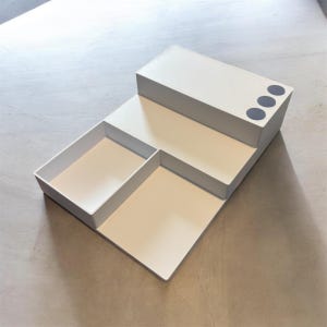 May include: A white, stepped desk organizer with three sections. The top section has three dark gray circular holes. The organizer is made of a smooth, matte material and is designed to hold office supplies.