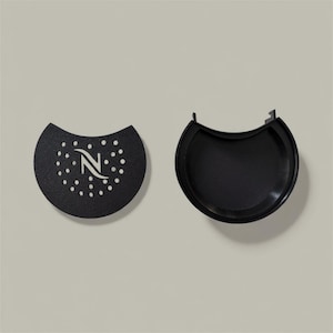 Puede incluir: Dos componentes negros y semicirculares para máquina de café. Uno tiene un diseño perforado con la letra "N" en el centro. El otro es una bandeja curva y sólida con una pequeña pestaña en un lado. Los artículos están sobre un fondo neutro.