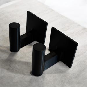 May include: Two black wall hooks. Each hook has a square backplate, a short horizontal arm, and a cylindrical hook. The hooks are made of a matte black material and are designed for mounting on a wall.