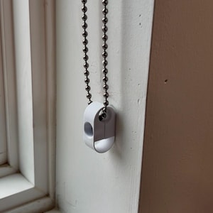 May include: A white plastic blind cord weight with a silver metal ball chain. The cord weight is oval-shaped with a hole in the center. The chain is hanging against a white wall.