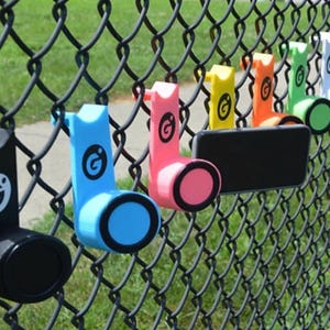 May include: A series of brightly coloured phone holders attached to a chain-link fence. The holders are in black, blue, pink, yellow, orange, green, white, and grey. Each has a circular base and a logo, designed for outdoor use.