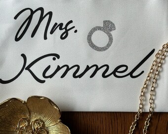 May include: A white canvas cosmetic bag with the text "Mrs. Kimmel" in black script and a silver glitter ring design. A gold-colored floral dish holds pearl earrings, and a gold chain is visible.