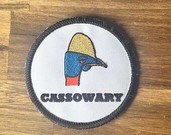 May include: A white, round, embroidered patch with a black border. The patch features a cartoon cassowary with a yellow helmet and the word "CASSOWARY" in black lettering.