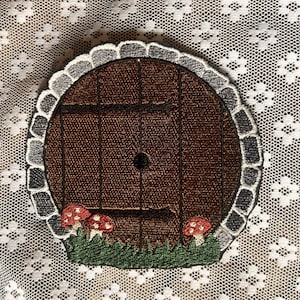 May include: Embroidered patch of a round wooden door with a stone frame, surrounded by green grass and red mushrooms.
