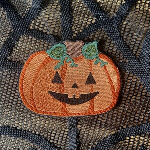 May include: An orange embroidered pumpkin patch with a carved jack-o'-lantern face. The pumpkin has a green stem and is on a black background.
