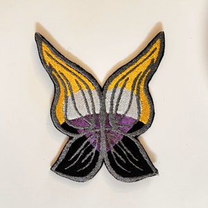May include: A yellow, white and purple butterfly-shaped embroidered patch. The butterfly wings are outlined in black and the centre of the butterfly is a purple triangle.