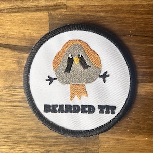 Könnte beinhalten: Ein runder weißer gestickter Aufnäher mit schwarzem Rand. Der Aufnäher zeigt einen Cartoon-Vogel mit grauem Körper, orangefarbenem Kopf und schwarzen Augen. Der Text "BEARDED TIT" ist in Schwarz unter dem Vogel gestickt.