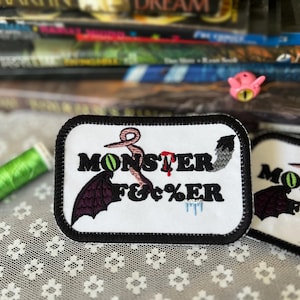 Puede incluir: Parche bordado con el texto "MONSTER F&c%ER" en negro, con un ojo verde, gotas de sangre rojas y una cola gris. El parche tiene un borde negro y está sobre un fondo blanco. Se ve una bobina de hilo verde.