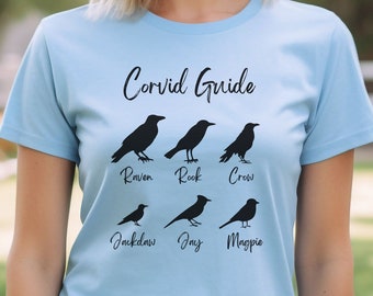 Op de afbeelding: Een lichtblauw T-shirt met een zwarte afbeelding van een gids voor kraaiachtigen. De afbeelding bevat silhouetten van zes verschillende vogels: raaf, roek, kraai, kauw, gaai en ekster. De tekst "Corvid Guide" is boven de vogels gedrukt.