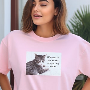May include: Pink t-shirt with a graphic of a cat and the text "life update: the voices are getting louder".