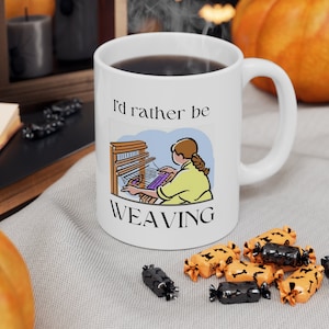 May include: White coffee mug with a black handle. The mug has a cartoon illustration of a person weaving on a loom. The text on the mug reads "I'd rather be weaving".