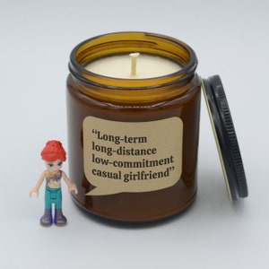 May include: A brown glass candle jar with a beige lid. The candle is lit and the jar has a brown paper label with the text "Long-term long-distance low-commitment casual girlfriend".