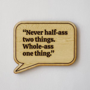May include: A wooden plaque in the shape of a speech bubble. The plaque has a light brown wood grain finish with a darker brown border. The text "Never half-ass two things. Whole-ass one thing." is engraved in a dark brown font.