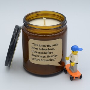 May include: A brown glass jar candle with a black lid, featuring the text "You know my code. Hoes before bros. Uteruses before duderuses. Ovaries before brovaries." A Lego figure on a skateboard is next to the candle.