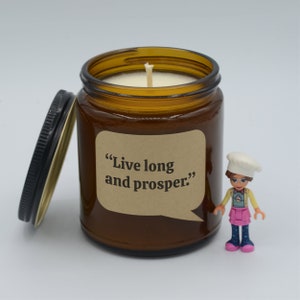 May include: A brown glass candle jar with a white candle and a brown paper label that says "Live long and prosper." The candle is next to a small plastic figurine of a girl wearing a white chef's hat and a pink dress.