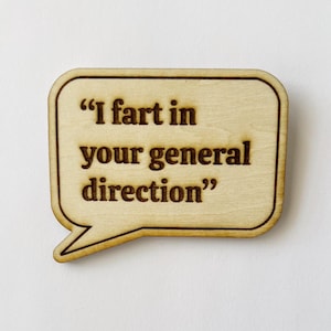 May include: A wooden pin in the shape of a speech bubble. The pin has a light wood color with a darker brown border. The text "I fart in your general direction" is engraved in the center of the pin.