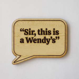 May include: A wooden sign in the shape of a speech bubble with the phrase "Sir, this is a Wendy's" in dark brown lettering. The sign has a light brown wood grain finish and a darker brown border.