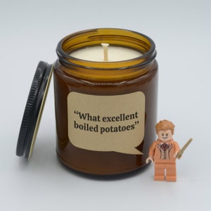 May include: A brown glass candle jar with a white candle inside. The jar has a brown paper label with the text "What excellent boiled potatoes". A small orange Lego figure with a wand is in front of the candle jar.